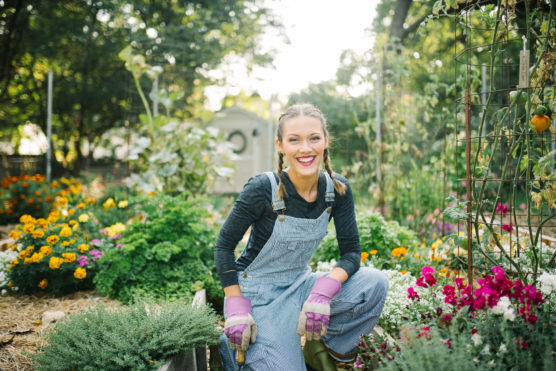 A woman wearing overalls and purple gloves kneels and smiles in a vibrant garden full of flowers and greenery, with sunlight filtering through the trees in the background. by Justin Salem Meyer