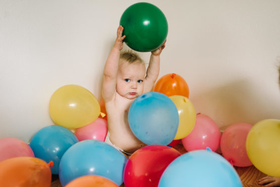 A baby sits surrounded by colorful balloons, holding a green balloon above their head with both hands, against a plain white wall. by Justin Salem Meyer