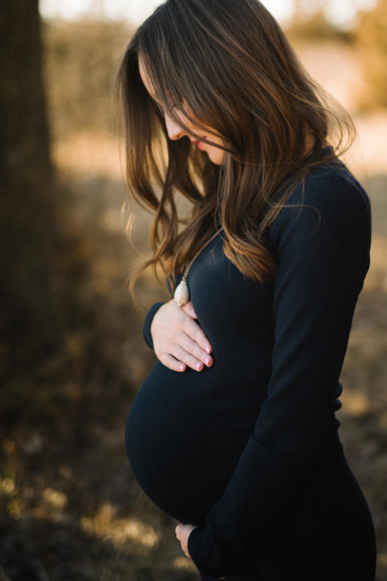 A woman with long brown hair stands outdoors, gently cradling her pregnant belly with both hands, wearing a long-sleeve black dress and looking down with a peaceful expression. by Justin Salem Meyer