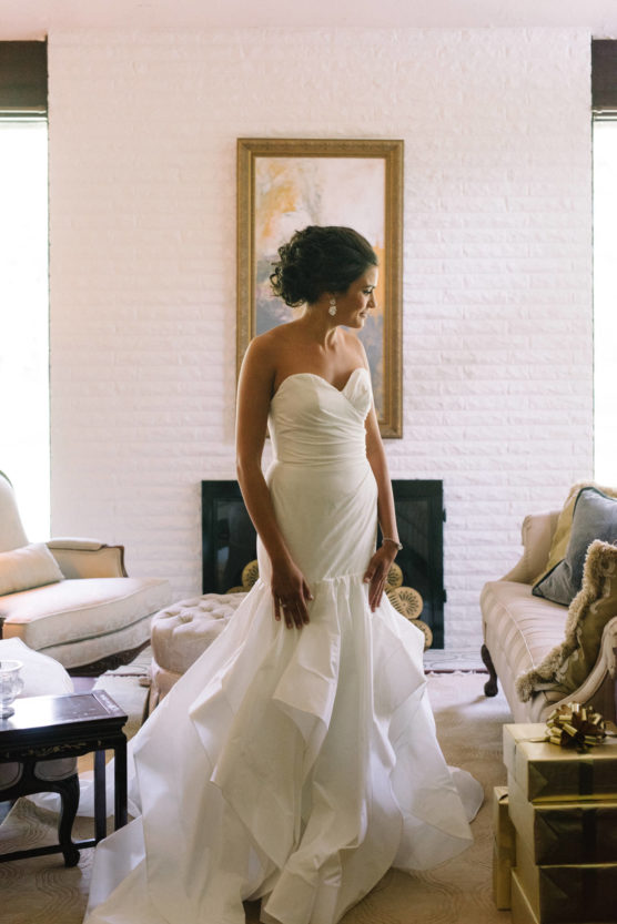 A woman in a strapless white wedding dress stands indoors, looking to the side. She has dark, curled hair styled up. The room has elegant furniture, a white brick fireplace, and a framed painting on the wall. by Justin Salem Meyer