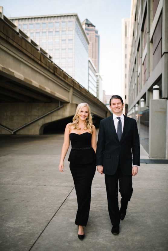 A woman and a man in formal black attire walk side by side on a city sidewalk, smiling. Tall buildings and a raised roadway are visible in the background. by Justin Salem Meyer