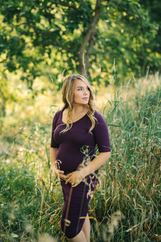 A woman in a fitted purple dress stands in tall grass and greenery, holding her midsection and looking to the side, with sunlit trees in the background. by Justin Salem Meyer