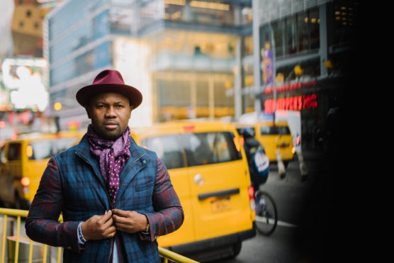 A man in a plaid jacket, purple scarf, and maroon hat stands on a city street with yellow taxis and blurred buildings in the background. by Justin Salem Meyer
