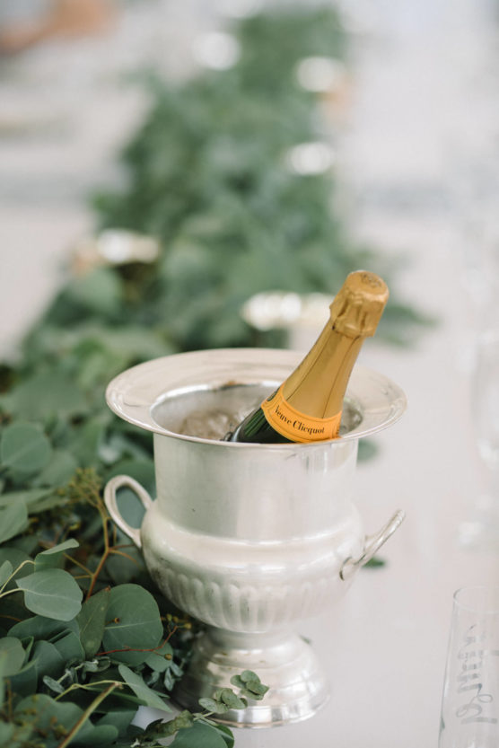 A bottle of Veuve Clicquot champagne in a silver ice bucket sits on a white table adorned with a green eucalyptus garland. by Justin Salem Meyer