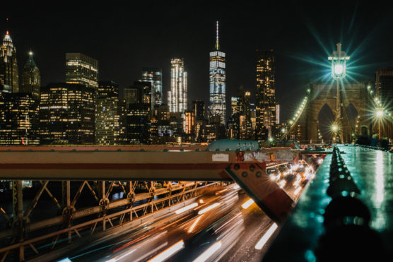 Night view of the New York City skyline with illuminated skyscrapers, including One World Trade Center, and blurred car lights on the Brooklyn Bridge in the foreground. by Justin Salem Meyer