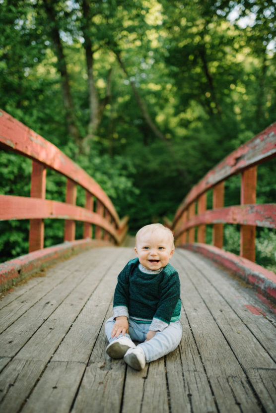 A smiling baby in a green sweater and light pants sits on a wooden bridge surrounded by lush green trees, with sunlight filtering through the leaves. by Justin Salem Meyer