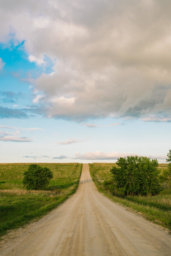 A long, straight dirt road stretches into the distance between grassy fields under a partly cloudy sky, with green trees on either side of the road and blue sky peeking through the clouds. by Justin Salem Meyer