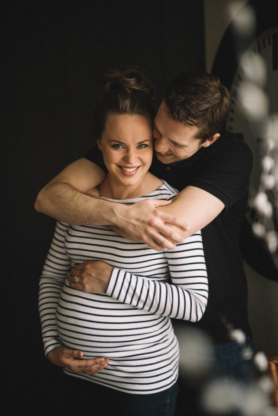 A smiling pregnant woman in a striped shirt is embraced from behind by a man in a black shirt. She holds her belly while he hugs her and leans in close, both appearing happy and affectionate. by Justin Salem Meyer