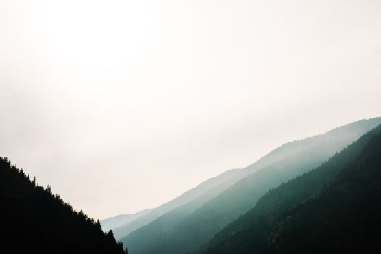 Soft sunlight shines through hazy clouds above a series of misty, forested mountains, creating a calm and tranquil landscape with gentle gradients of blue and green. by Justin Salem Meyer