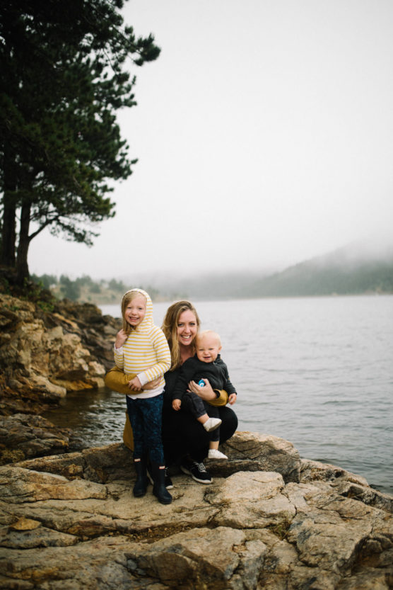 A woman and two young children sit on rocky ground by a lake, surrounded by trees and misty mountains. The woman and children are smiling, dressed in warm clothes, with water and foggy scenery in the background. by Justin Salem Meyer