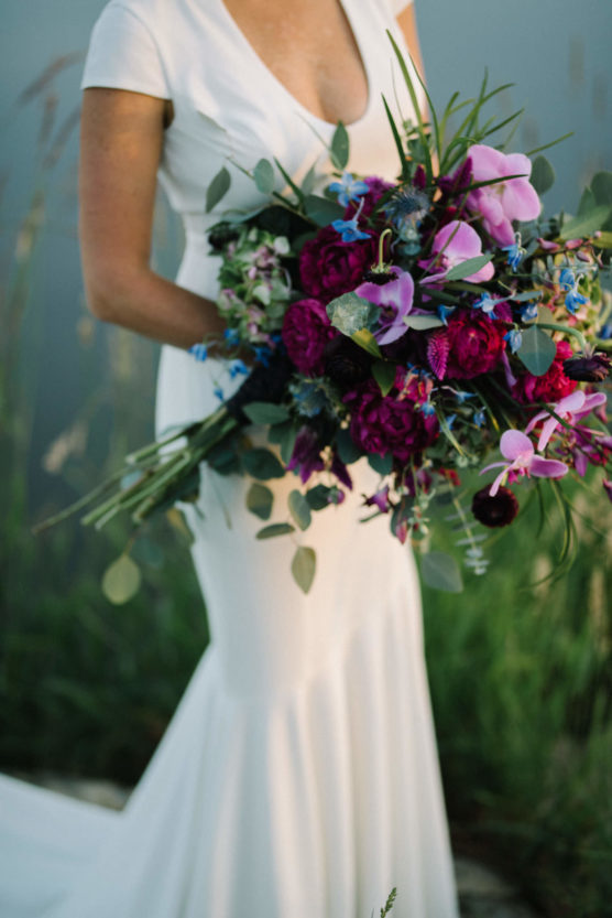 A bride in a white dress holds a large, colorful bouquet with deep pink, purple, and blue flowers and greenery, standing outdoors with blurred green grass in the background. by Justin Salem Meyer