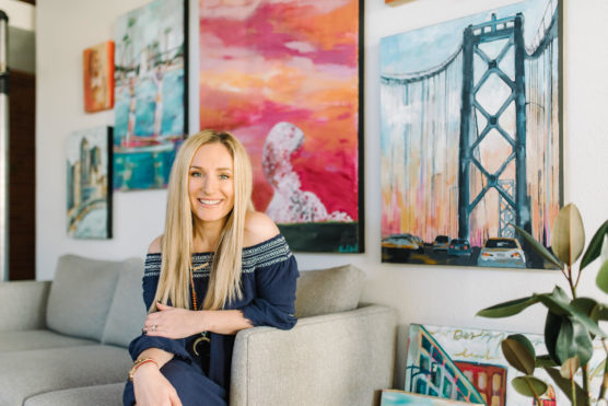A woman with long blonde hair sits on a beige couch, smiling. Behind her, colorful paintings decorate a white wall, including a bridge and sunset scene. A plant is visible in the foreground. by Justin Salem Meyer