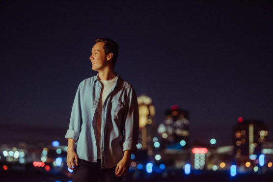A young man in a light blue shirt stands smiling at night with a city skyline and colorful lights blurred in the background. by Justin Salem Meyer