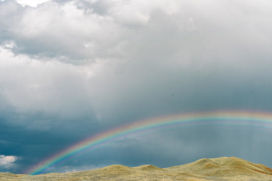 A faint rainbow arcs above rolling hills under a cloudy, overcast sky, with patches of sunlight highlighting parts of the landscape. by Justin Salem Meyer