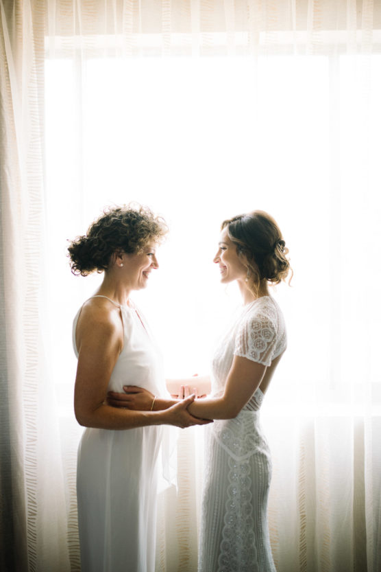 Two women in white dresses stand facing each other, holding hands and smiling, with soft natural light streaming through sheer curtains behind them. by Justin Salem Meyer