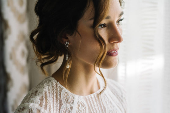 A woman in a white lace dress with loose, wavy hair gazes thoughtfully out of a softly lit window. She wears pearl earrings and natural makeup, with delicate strands of hair framing her face. by Justin Salem Meyer