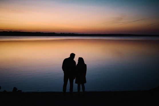 Silhouette of two people standing close together by a calm lake at sunset, with a colorful sky reflecting on the water. by Justin Salem Meyer