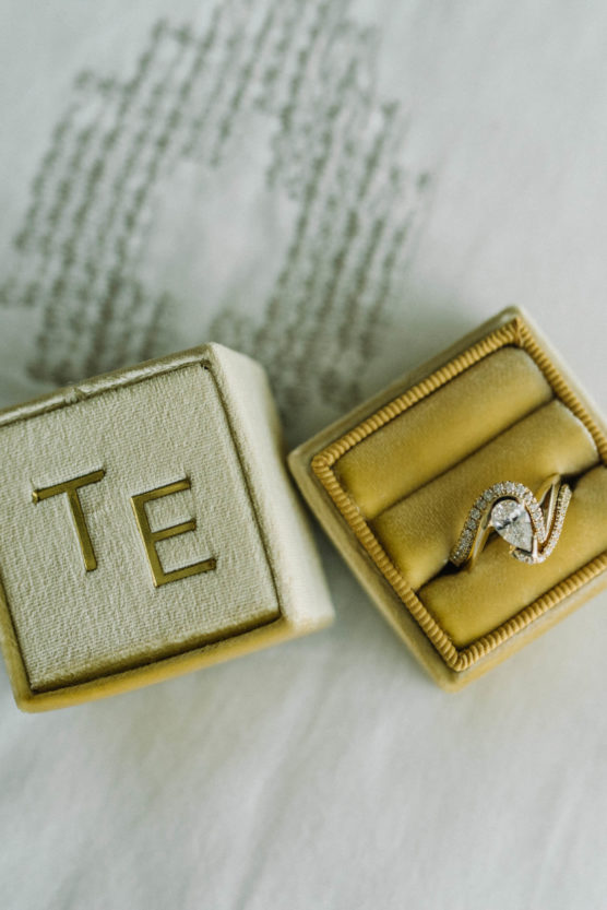 A diamond engagement ring with a teardrop-shaped stone and a twisted band rests in a beige velvet ring box. Another matching box with the initials TE is placed beside it on a soft, light surface. by Justin Salem Meyer