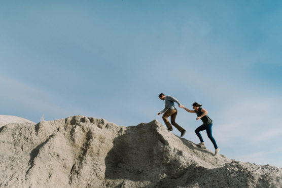 Two people holding hands climb up a rocky, sandy hill under a clear blue sky, with one person leading the other as they move upwards together. by Justin Salem Meyer