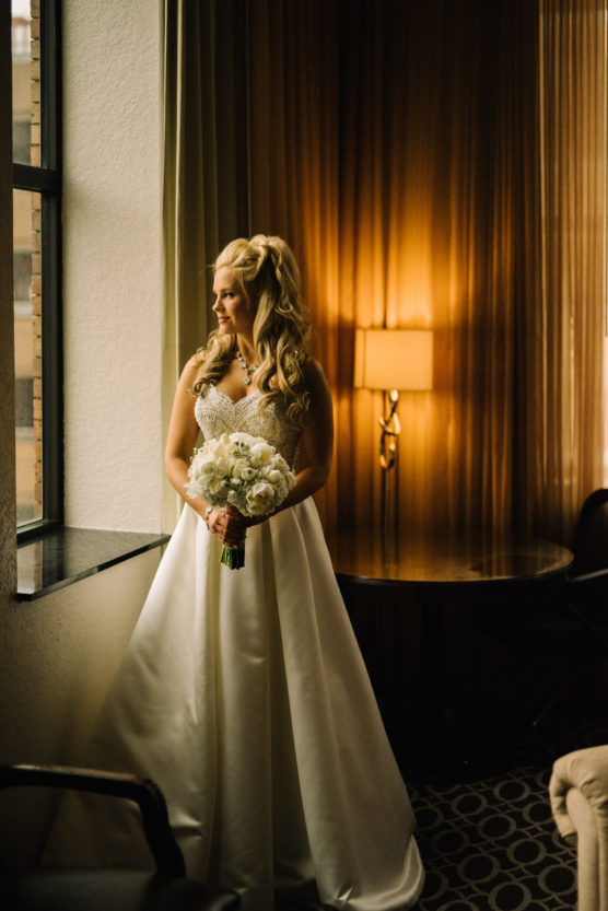 A bride in a white gown holding a bouquet of white flowers stands by a window, looking outside. Warm light from a lamp illuminates the room, creating a soft, elegant atmosphere. by Justin Salem Meyer