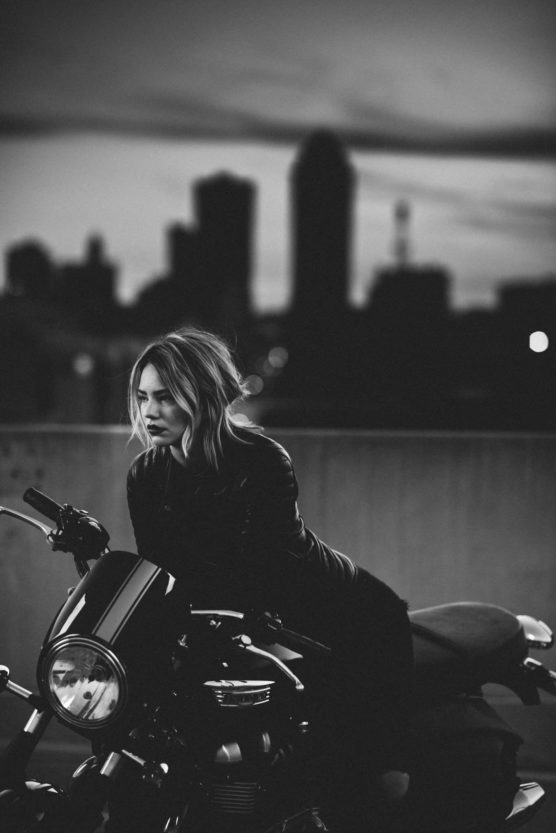A woman with wavy hair sits on a parked motorcycle, gazing pensively into the distance. The background features a city skyline at dusk, with buildings silhouetted against a darkening sky. The image is in black and white. by Justin Salem Meyer
