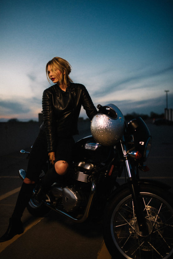 A woman in a black leather jacket and ripped jeans sits on a motorcycle at dusk, with a silver helmet resting on the handlebars and a dramatic sky in the background. by Justin Salem Meyer