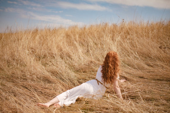A woman with long red hair, wearing a white dress, sits on dry, golden grass in a field, facing away and looking toward the sky on a partly cloudy day. by Justin Salem Meyer