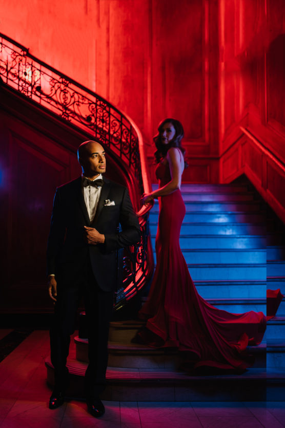 A man in a black tuxedo stands at the base of a grand staircase, while a woman in a long, elegant red gown poses on the steps. The scene is dramatically lit with red and blue lighting. by Justin Salem Meyer