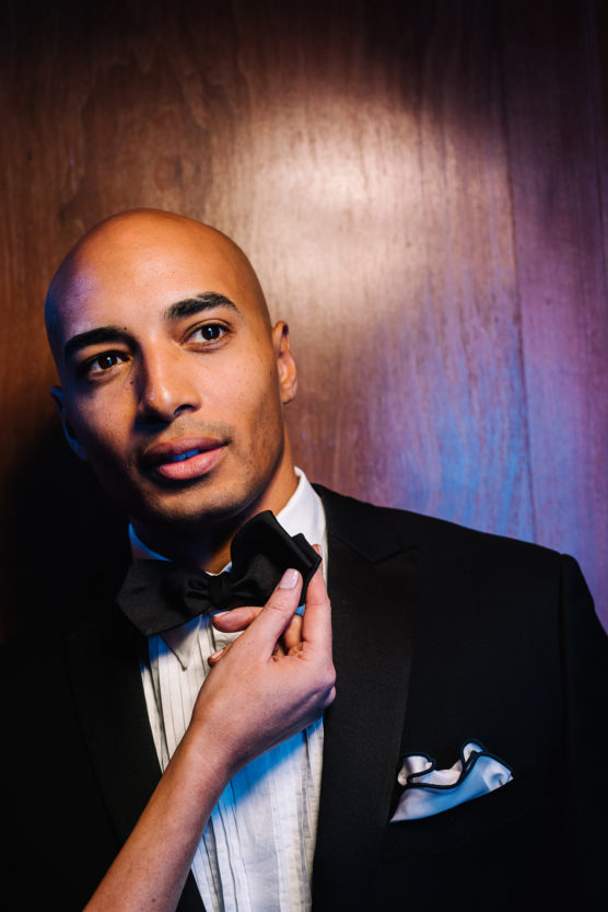 A man in a tuxedo stands against a wooden background while a hand adjusts his bow tie. He looks slightly to the side, with dramatic lighting highlighting his face and outfit. by Justin Salem Meyer