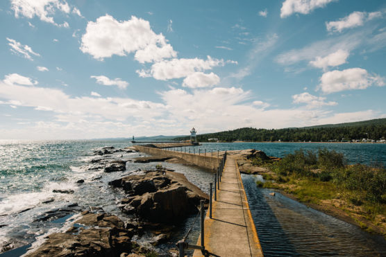 A concrete pier stretches out into a large, blue lake with a lighthouse at the end. Rocky shoreline and green vegetation line the water under a bright, partly cloudy sky. by Justin Salem Meyer