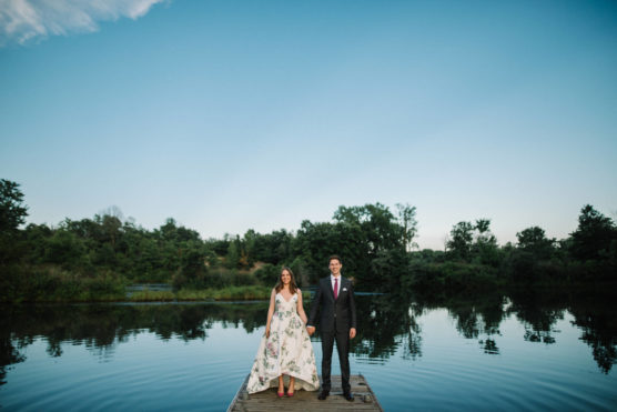 A woman in a white floral dress and a man in a dark suit stand side by side on a wooden dock over a calm lake, with trees and a clear blue sky in the background. by Justin Salem Meyer