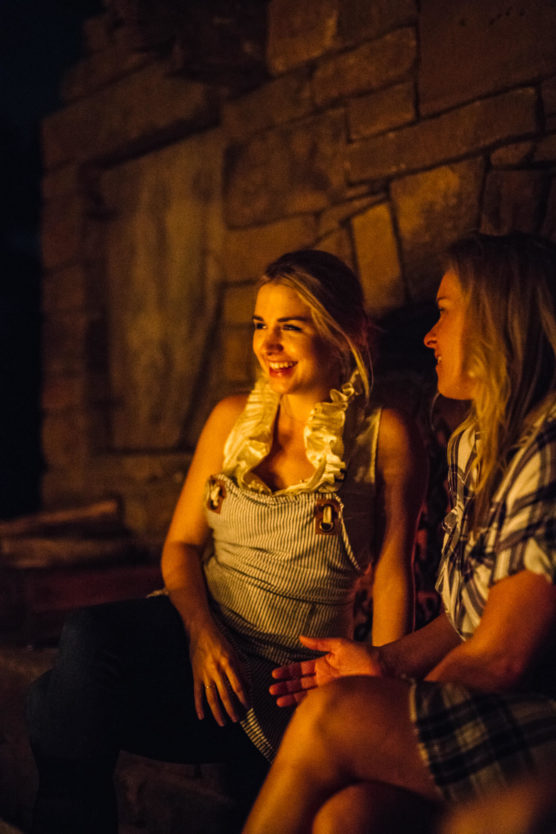 Two women sit together by a stone fireplace at night, warmly lit by firelight. One woman smiles brightly while the other gestures as they enjoy a relaxed conversation. by Justin Salem Meyer