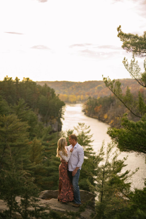 A couple stands on a rocky overlook surrounded by trees, embracing and smiling at each other, with a river and forested hills in the background under a soft, golden sky. by Justin Salem Meyer