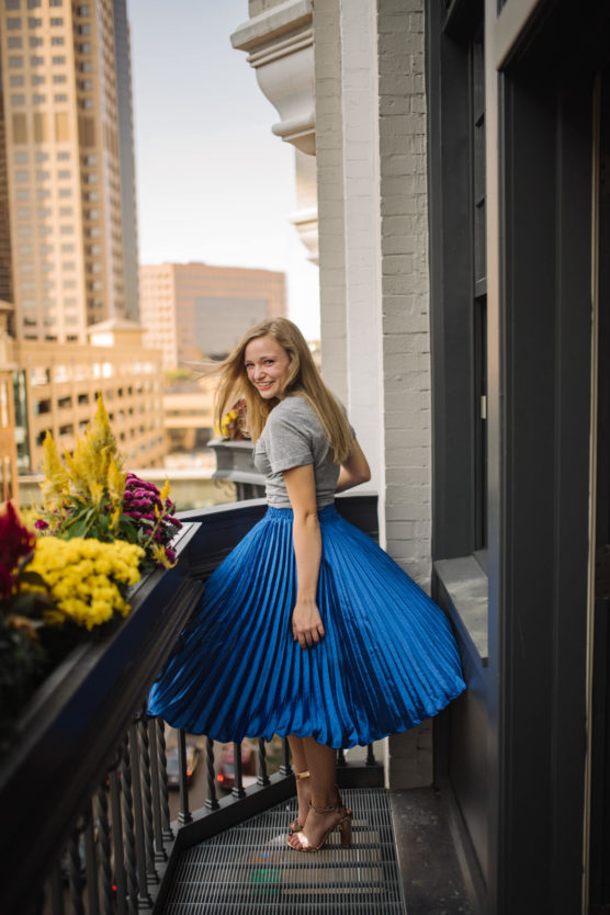 A woman with long blonde hair, wearing a grey top and a bright blue pleated skirt, smiles while twirling on a city balcony decorated with yellow and red flowers. Tall buildings are visible in the background. by Justin Salem Meyer