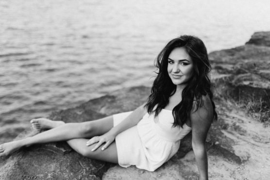 A young woman in a white dress sits on a rock by the water, looking at the camera and smiling. The image is in black and white. by Justin Salem Meyer