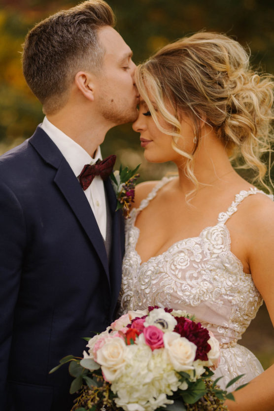 A groom in a dark suit kisses the forehead of his bride, who wears a white lace dress and holds a bouquet of roses. They stand close together outdoors, sharing an intimate moment. by Justin Salem Meyer