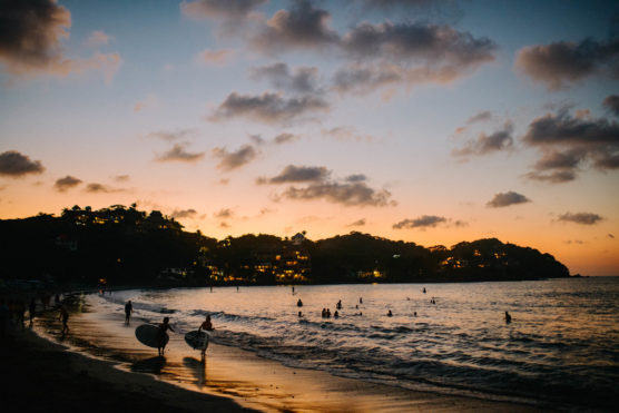 People swim and surf at a beach during sunset. The sky is filled with clouds, and lights from hillside buildings illuminate the background, creating a peaceful and lively seaside scene. by Justin Salem Meyer