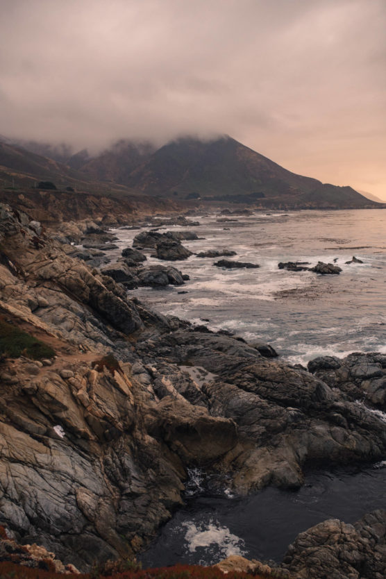 Rocky coastline with waves crashing against the shore, under a cloudy sky. Large mountains in the background are partially covered by mist. The scene appears moody and atmospheric, suggesting an overcast day. by Justin Salem Meyer