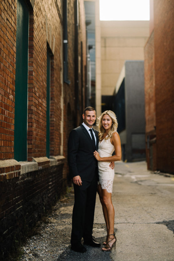 A man in a black suit and tie stands next to a woman in a short white lace dress and heels. They smile and pose together in a narrow, sunlit alleyway between brick buildings. by Justin Salem Meyer