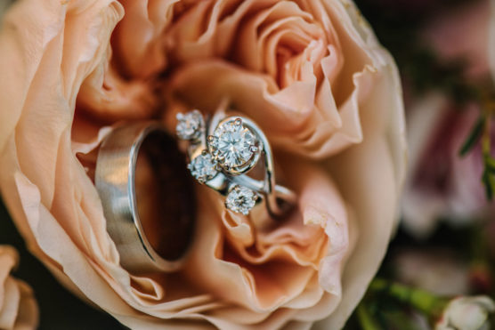Close-up of a diamond engagement ring, a wedding band, and another diamond ring nestled inside the petals of a peach-colored rose. by Justin Salem Meyer