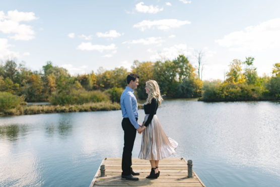 A couple stands holding hands at the end of a wooden dock on a calm lake, surrounded by trees with autumn foliage under a bright, partly cloudy sky. The womans skirt flows in the breeze. by Justin Salem Meyer