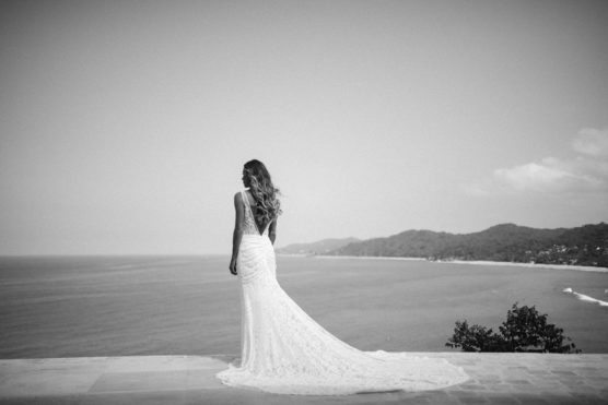 A bride in a long, lace wedding gown stands on a stone terrace, gazing out over the ocean and distant coastline under a clear sky. The distant hills and water create a serene, scenic backdrop. by Justin Salem Meyer