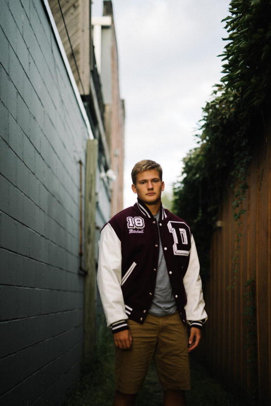 A young man stands in a narrow alleyway wearing a maroon and white varsity jacket with 18 and Mitchell on it, over a gray shirt and khaki shorts, framed by fences and greenery. by Justin Salem Meyer