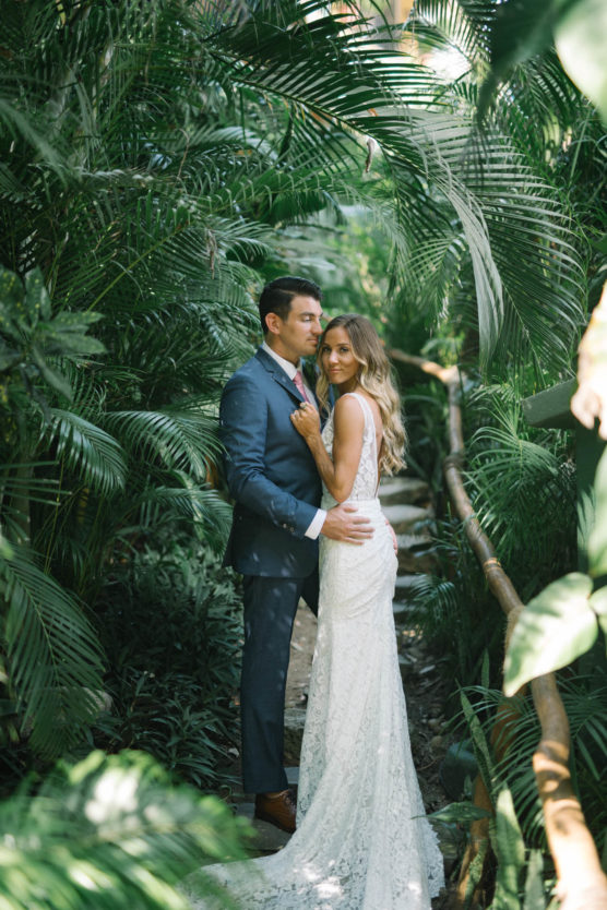 A bride in a white lace gown and a groom in a blue suit embrace on a lush, tropical garden path surrounded by green foliage, looking serene and romantic. by Justin Salem Meyer