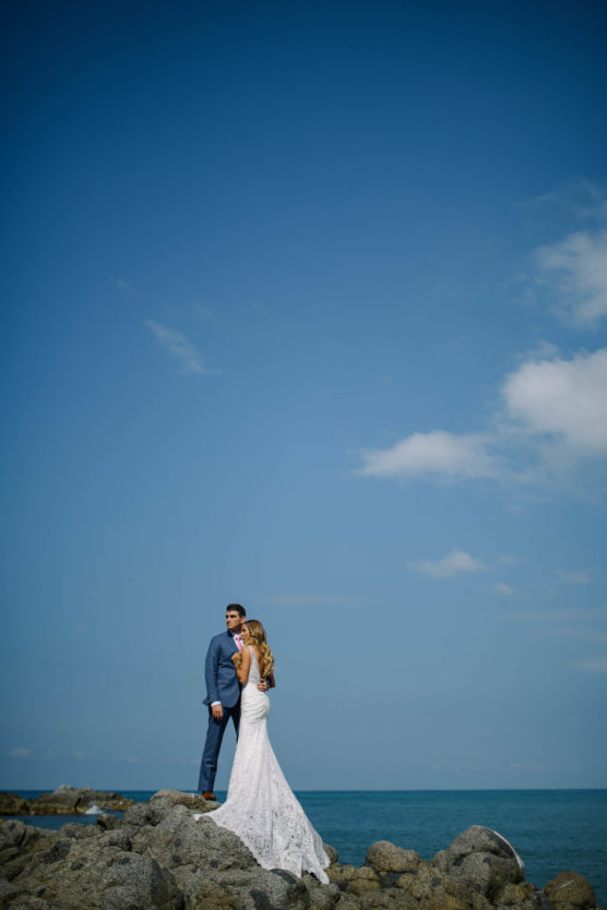 A bride in a white dress and a groom in a blue suit stand together on rocky shore under a clear blue sky, with the ocean in the background. by Justin Salem Meyer