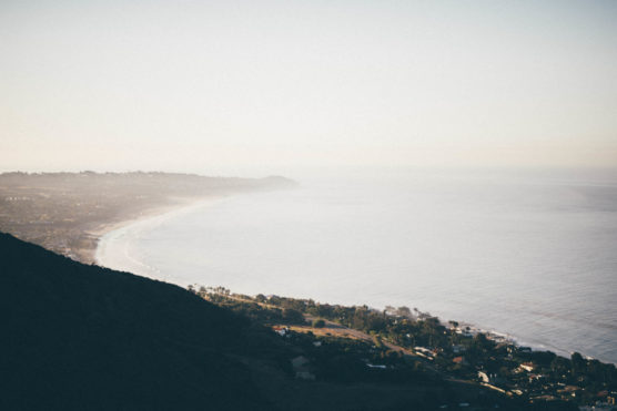 A hazy coastal landscape with gentle waves meeting a long, curving shoreline. A hillside in the foreground overlooks houses and greenery along the coast, under a pale sky. by Justin Salem Meyer
