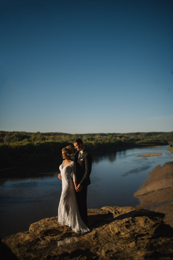A bride and groom stand on a rocky ledge by a river, holding hands and looking into the distance under a clear blue sky, with green trees and water in the background. by Justin Salem Meyer