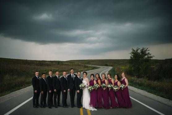 A wedding party stands together on a rural road under a cloudy sky. The bride and groom are surrounded by bridesmaids in burgundy dresses holding bouquets and groomsmen in black suits. The road winds into the distance through grassy fields. by Justin Salem Meyer