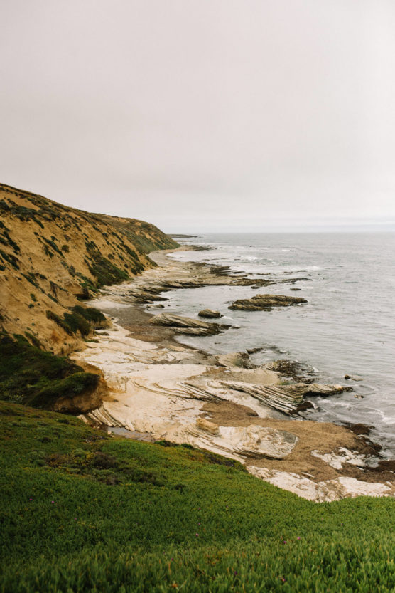A coastal landscape with grassy cliffs sloping down to rocky, beige shoreline and the ocean under an overcast sky. Waves gently wash over the rocks, and sparse vegetation covers parts of the cliffs. by Justin Salem Meyer