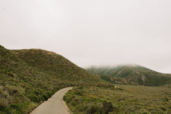 A narrow paved road winds through green hills under an overcast sky, with low clouds partially covering the distant mountains. by Justin Salem Meyer